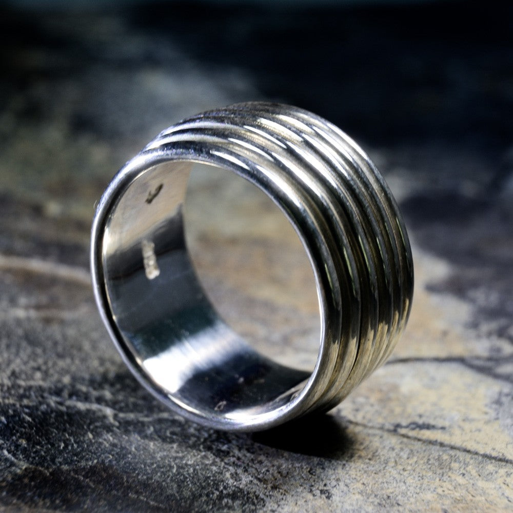 A silver-toned ring with a wide, horizontally grooved band, photographed on a dark textured surface with a shallow depth of field.