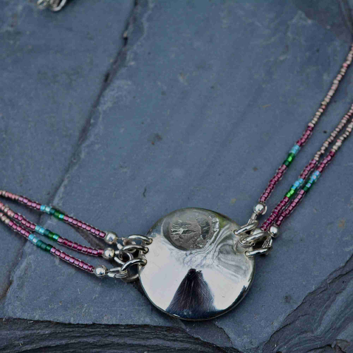 A silver-toned disc pendant necklace with a coin core, hanging from three beaded strands in pink, green, and blue on a dark slate background.