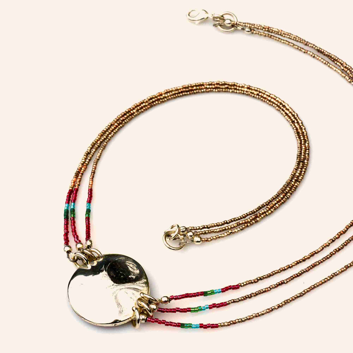 A multi-strand beaded necklace with a polished silver disc pendant, featuring gold, bronze, and red seed beads with small turquoise accents, displayed on a light cream background.