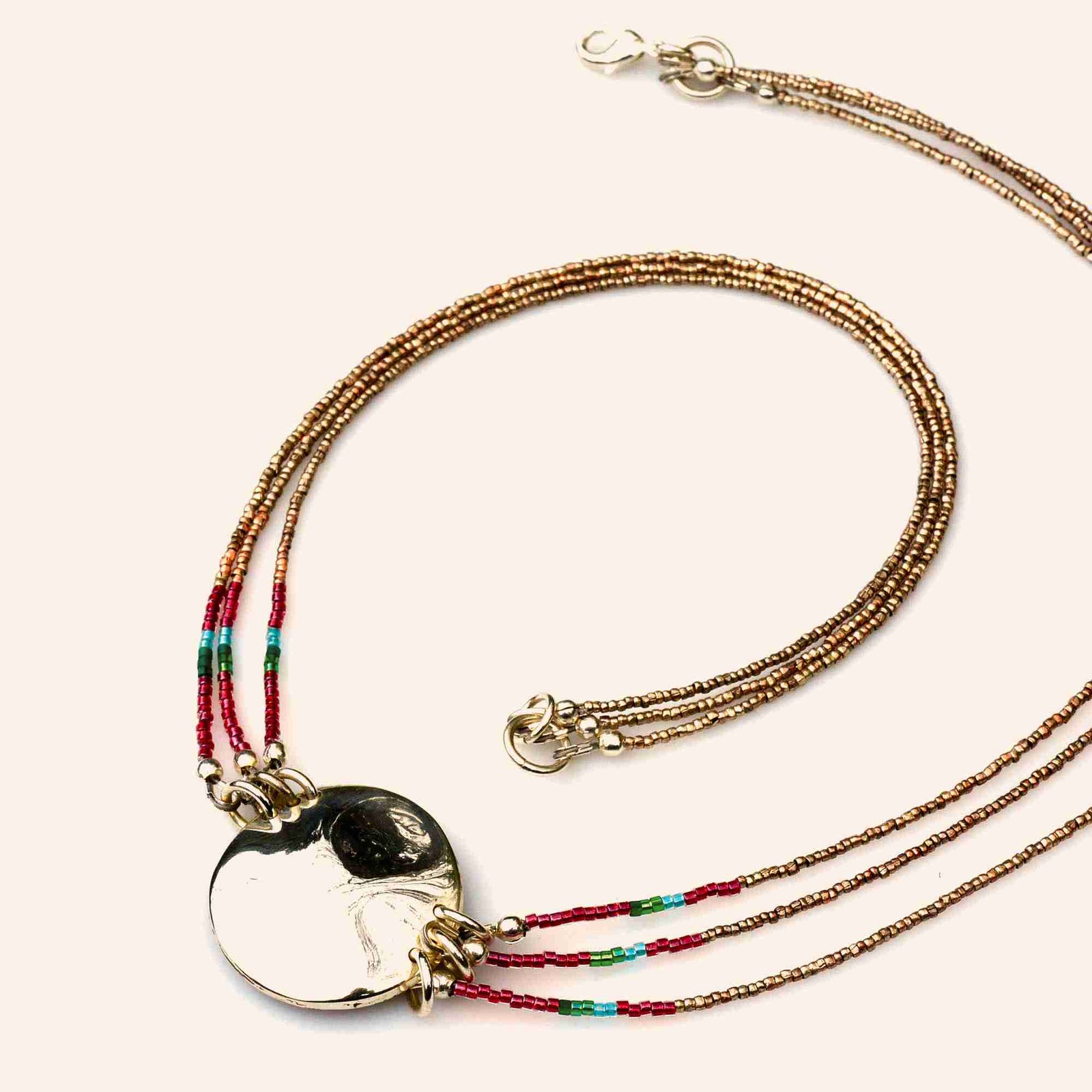 A silver-toned pendant necklace with a polished oval medallion, suspended from multiple delicate strands of small beads in red, green, turquoise, and cream, resting on a dark slate surface.