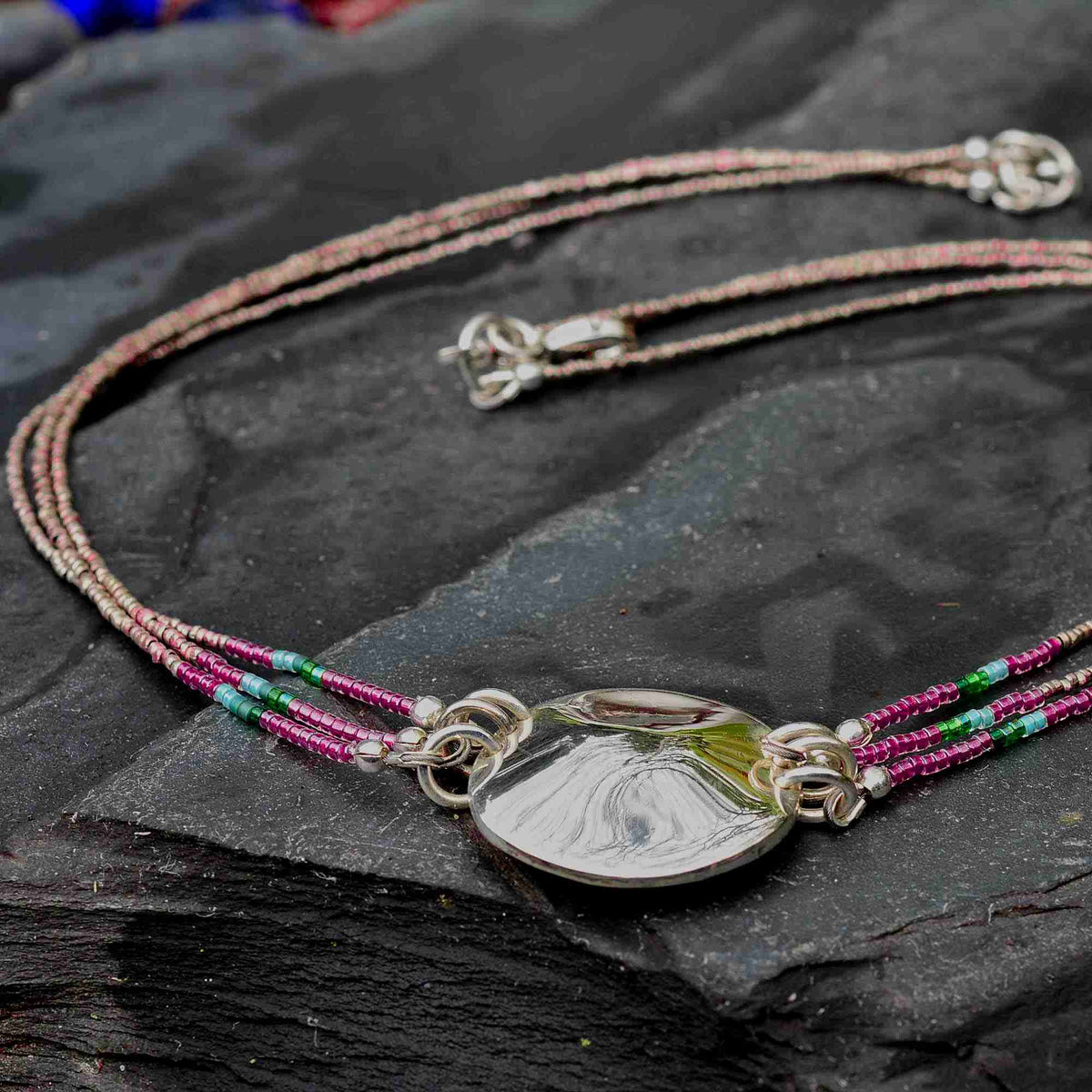 A close-up of a multi-strand beaded necklace with a polished silver pendant, displayed on a dark textured slate surface.