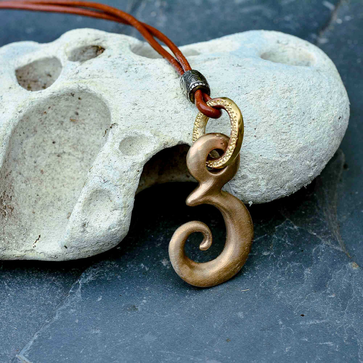 A close-up of a bronze-colored spiral pendant on a brown cord, resting against a white textured stone on a dark slate surface.
