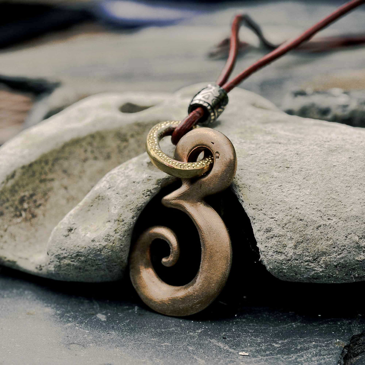 A close-up of a carved spiral pendant necklace in a warm metallic finish, hanging from a brown cord with a silver-toned bead, resting against a gray stone.