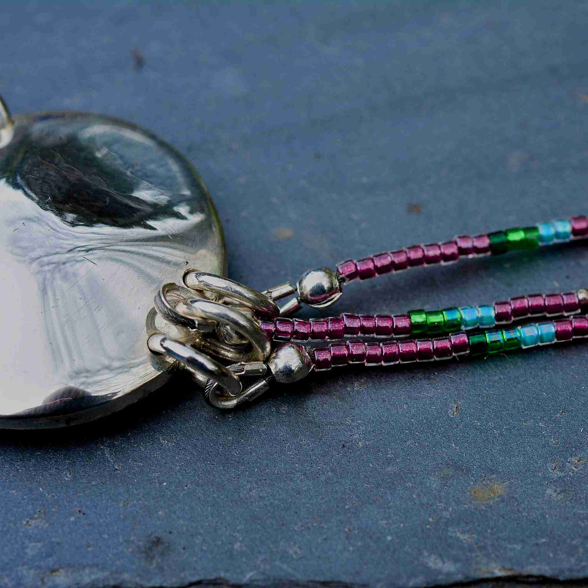 Close-up of a silver pendant necklace with a polished silver heart-shaped pendant and multi-colored seed beads in pink, green, and blue on a dark slate background.