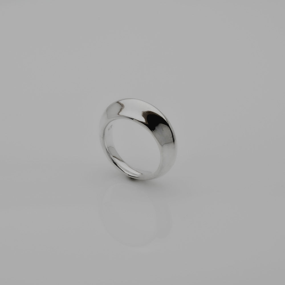 A polished silver band ring standing upright on a smooth light gray surface with a soft reflection below.