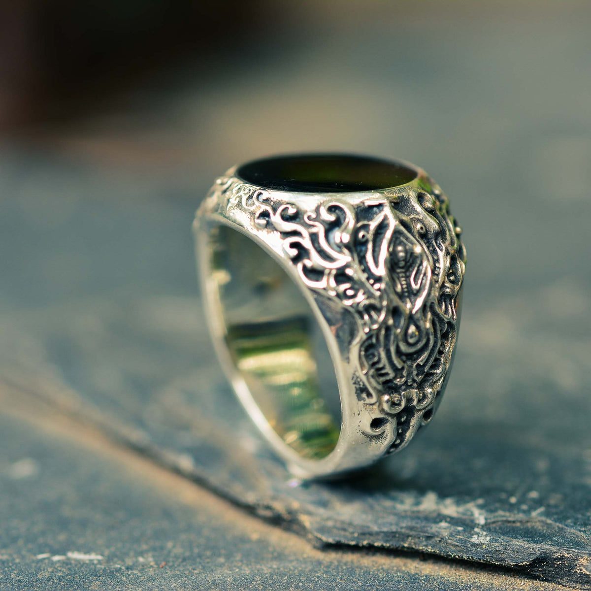 A large silver ring with intricate textures and a polished onyx color resin surface, displayed on a stone surface.