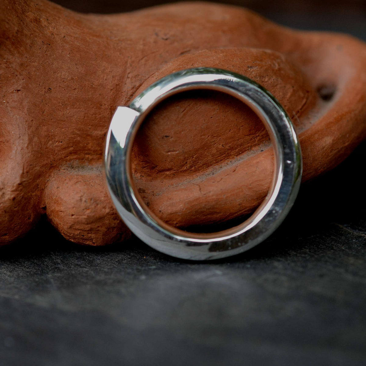 A polished silver-tone metal ring with a wide, flat circular face and a small triangular notch on one side, resting against a textured terracotta clay prop on a dark surface.