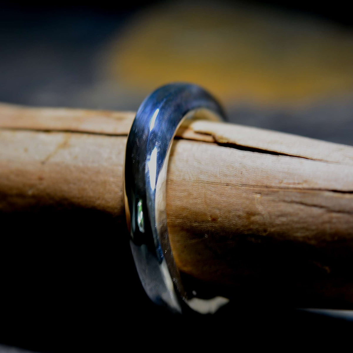 A polished dark metallic ring band, appearing black or gunmetal gray, resting on a piece of natural wood against a dark, blurred background.