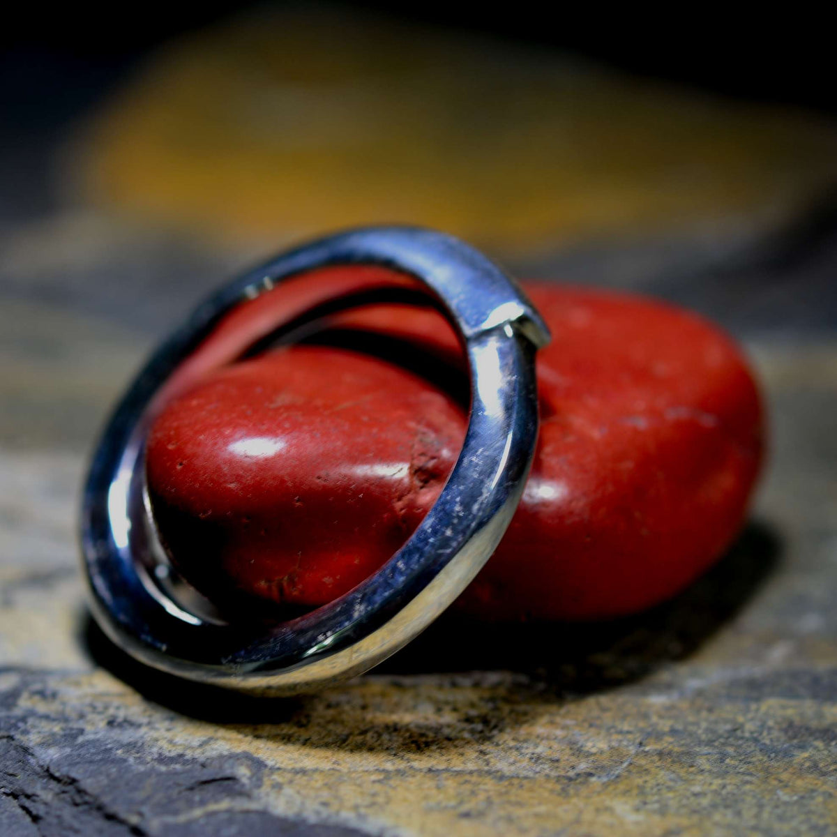 A polished silver-toned metal ring with a unique double-loop or interlocking design, resting on a textured dark stone surface with a smooth red stone behind it.