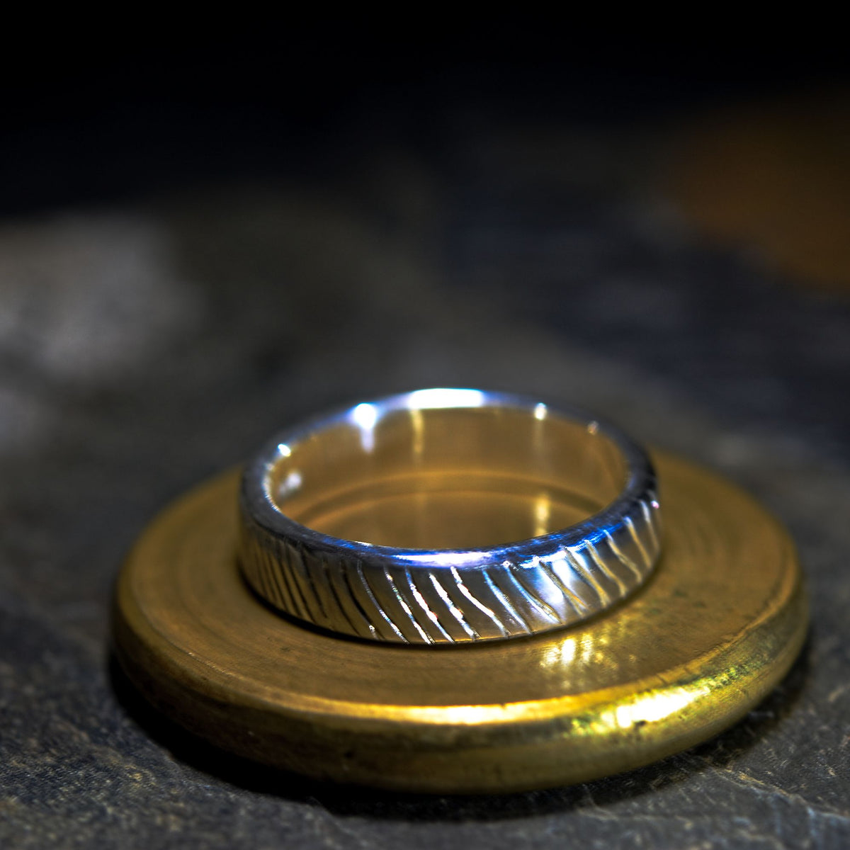 A textured silver band ring resting on a round gold-colored metal stand against a dark, blurred background.