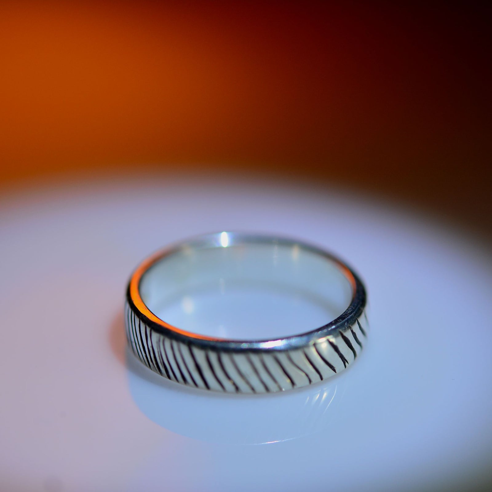 A silver band ring with a textured, wavy linear pattern on its outer surface, resting on a reflective white surface with a warm orange gradient background.