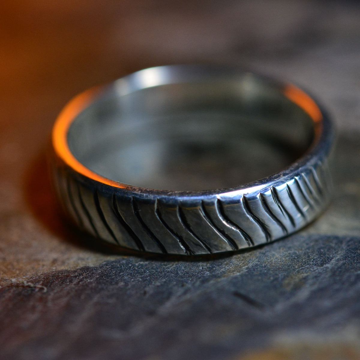 A close-up of a textured silver ring with a wavy, grooved pattern, resting on a dark, rustic stone surface.