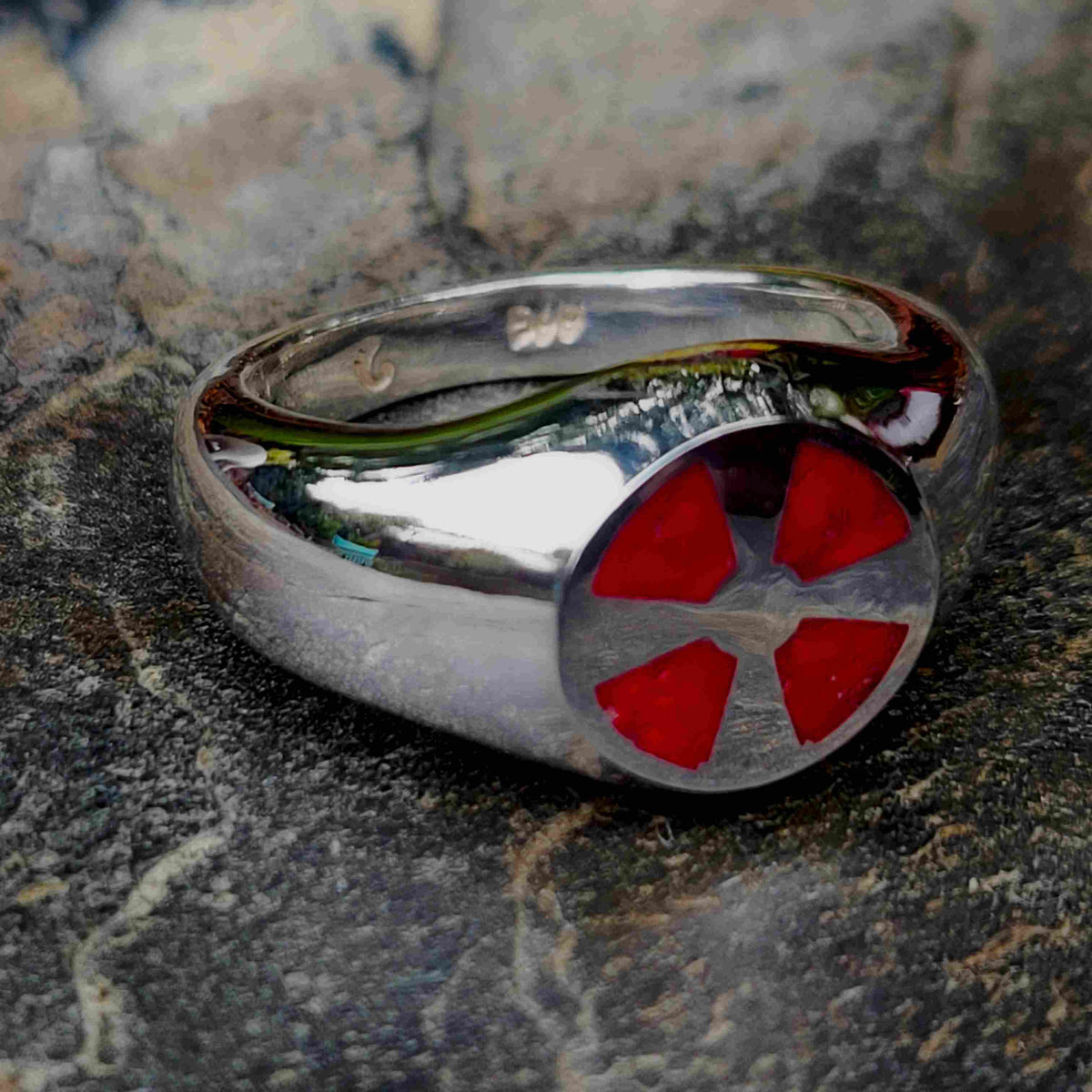 A polished silver ring featuring a circular face with four red triangular inlays arranged in a cross-like pattern, resting on a textured dark stone surface.