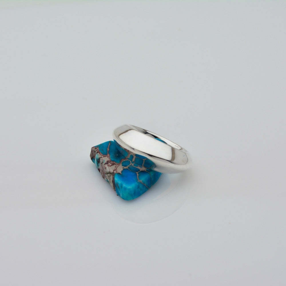 A sterling silver ring with a wide, polished band, featuring a large, raw turquoise stone with brown matrix veining, set against a plain light gray background.