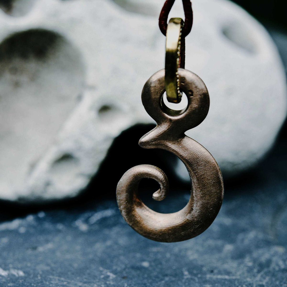 A close-up of a dark metallic pendant shaped like the number 8, hanging from a dark cord against a blurred gray stone and blue surface background.