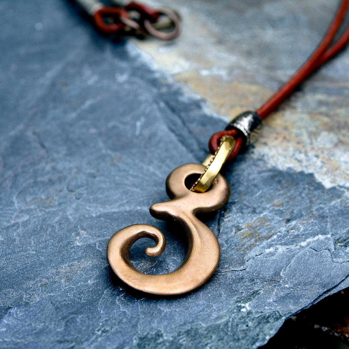 A close-up of a metallic pendant necklace on a dark slate surface. The pendant is a stylized number 3 in a warm copper or bronze tone, attached to a red cord with a small silver spacer.