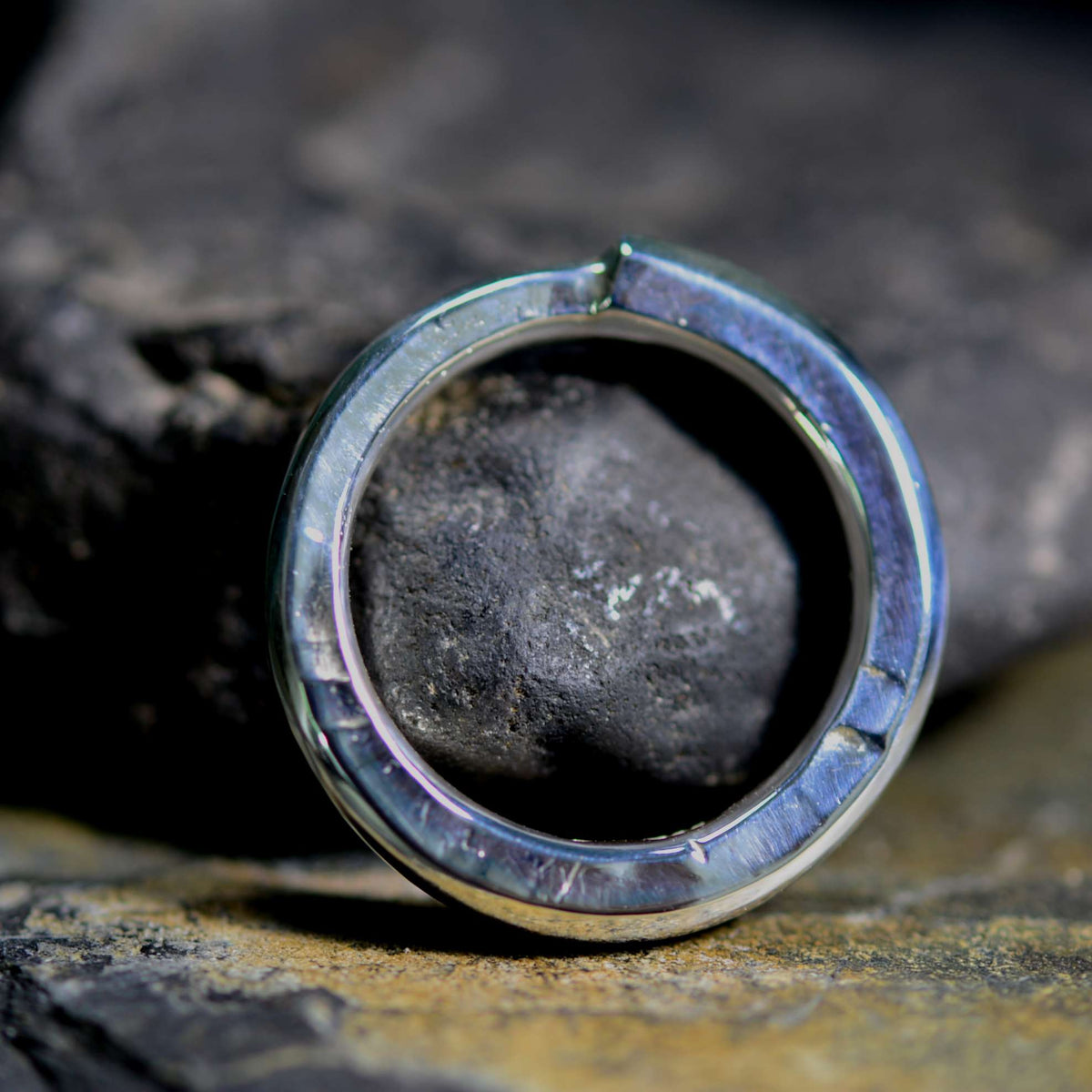 A silver-toned ring with a circular inlay of iridescent blue-purple material, holding a dark stone, photographed close-up on a textured rock surface.