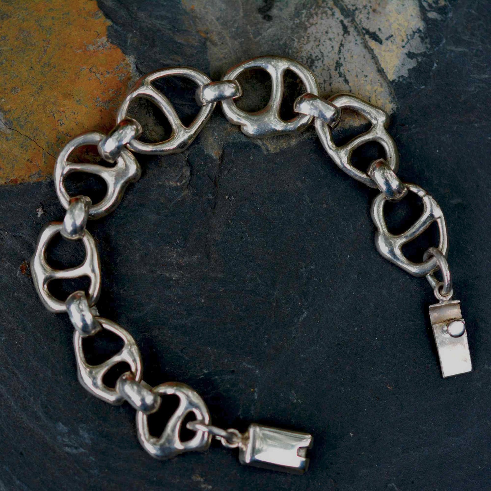 A silver-tone chain bracelet made of interlocking pull-tab style links, photographed on a dark textured surface with a yellowish stone patch in the upper left.