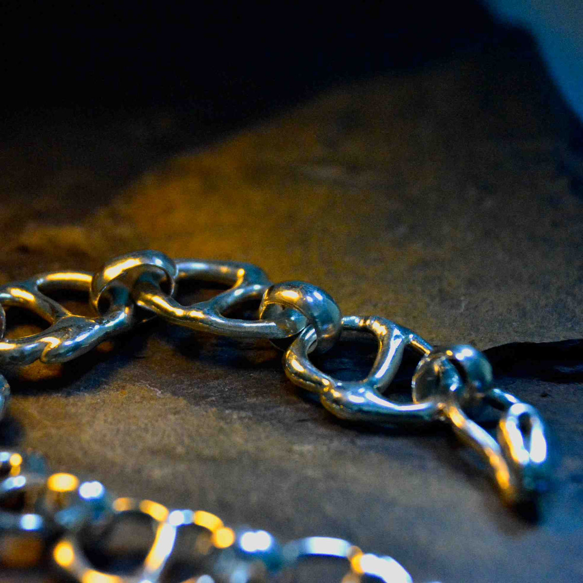 A close-up of a chunky silver chain with thick interlocking links resting on a dark textured surface under dramatic warm and cool lighting.