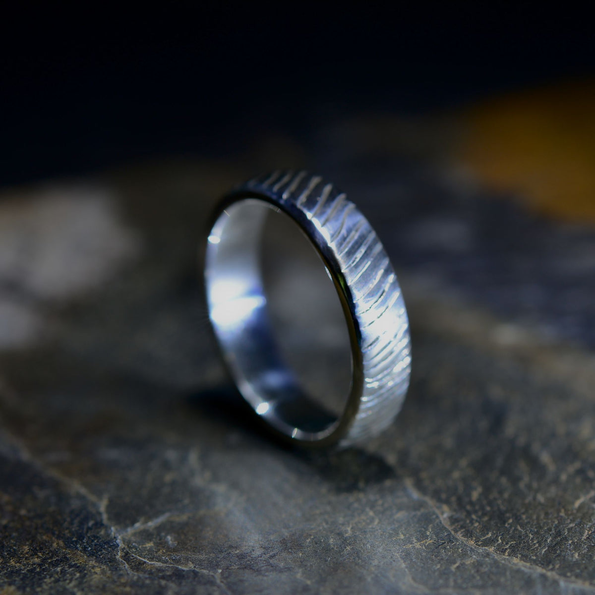 A textured silver band ring with a brushed, ribbed exterior and a polished interior, resting on a dark, textured stone surface.