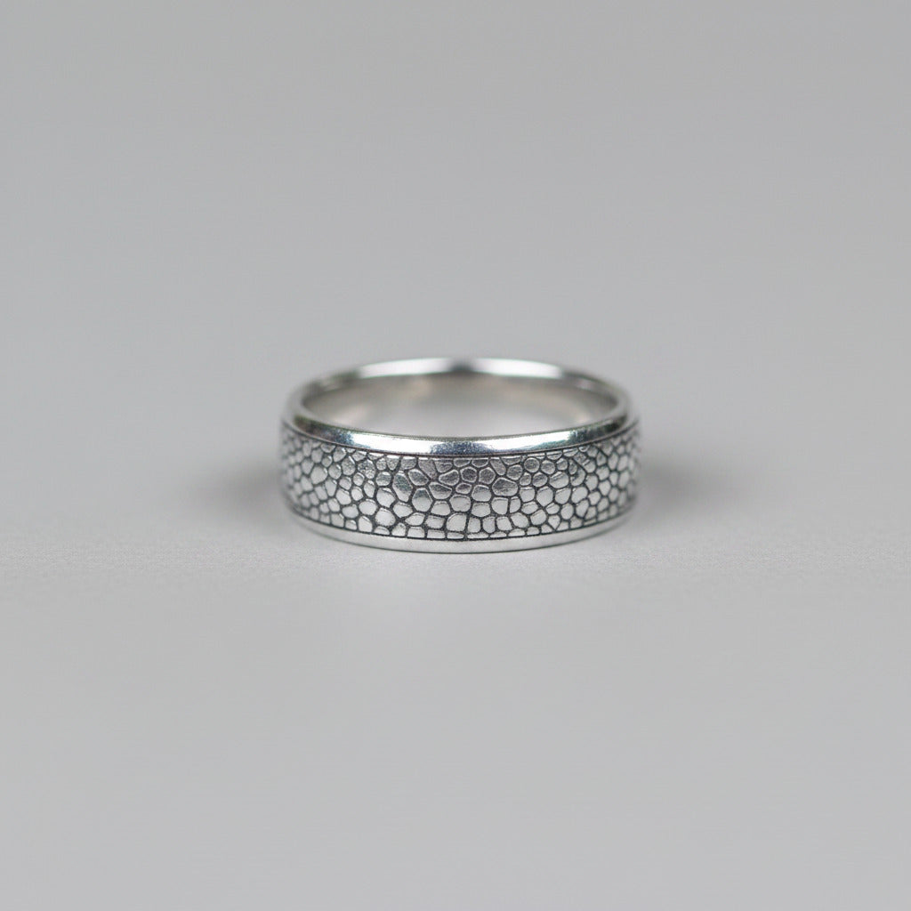 A silver band ring with a textured, raised honeycomb or scales-like pattern on the front, photographed on a neutral light gray background.
