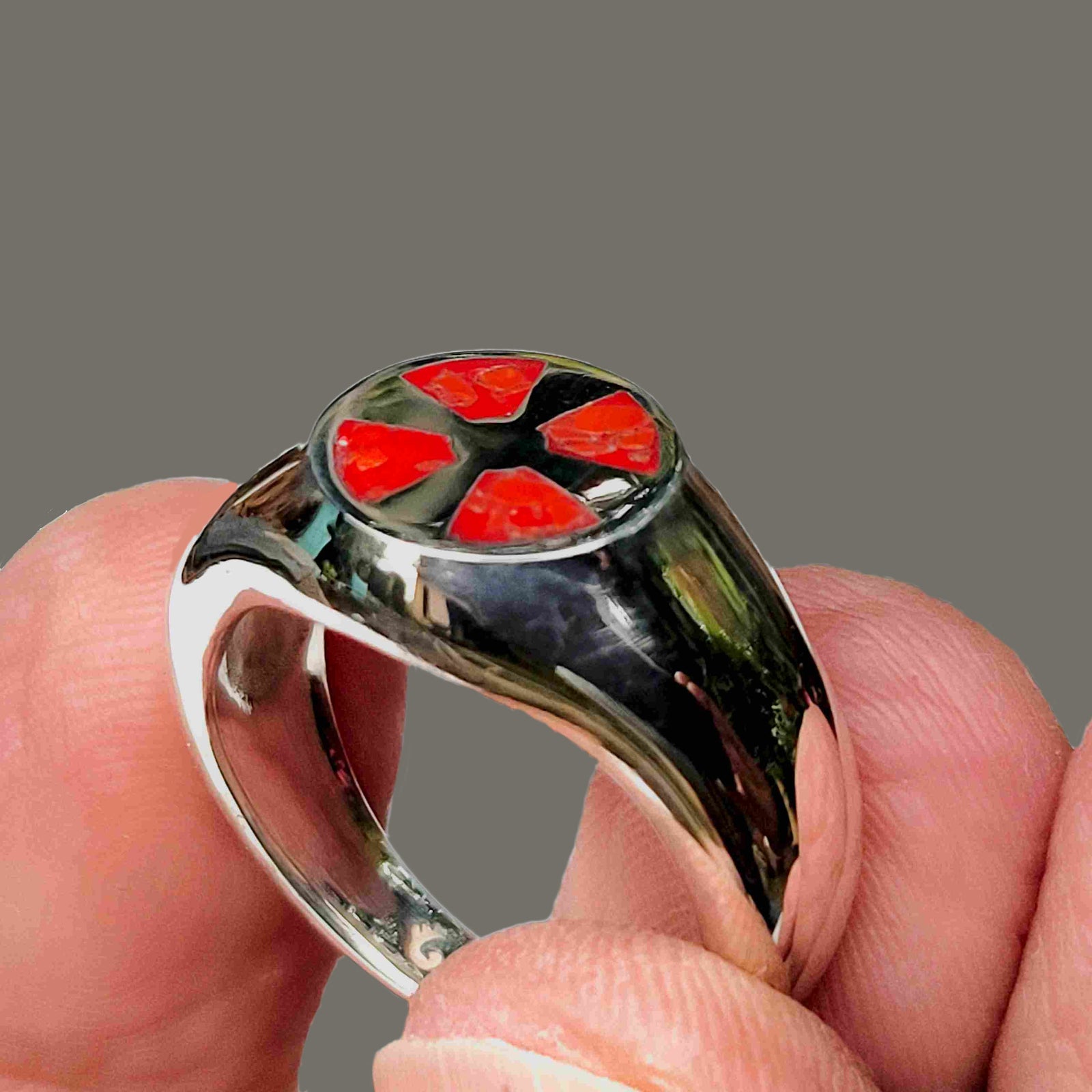 A close-up of a hand holding a polished silver signet ring with a red enamel inlay in the shape of a cross.