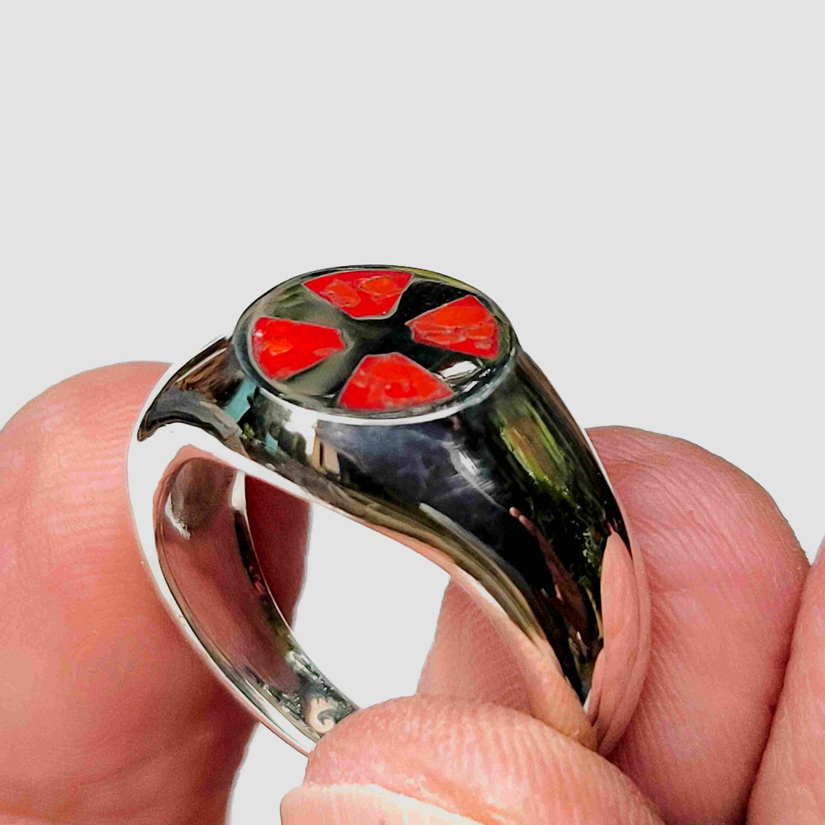 A polished silver ring held in a hand, featuring a circular top with four bright red petal-like inlays on a black background.