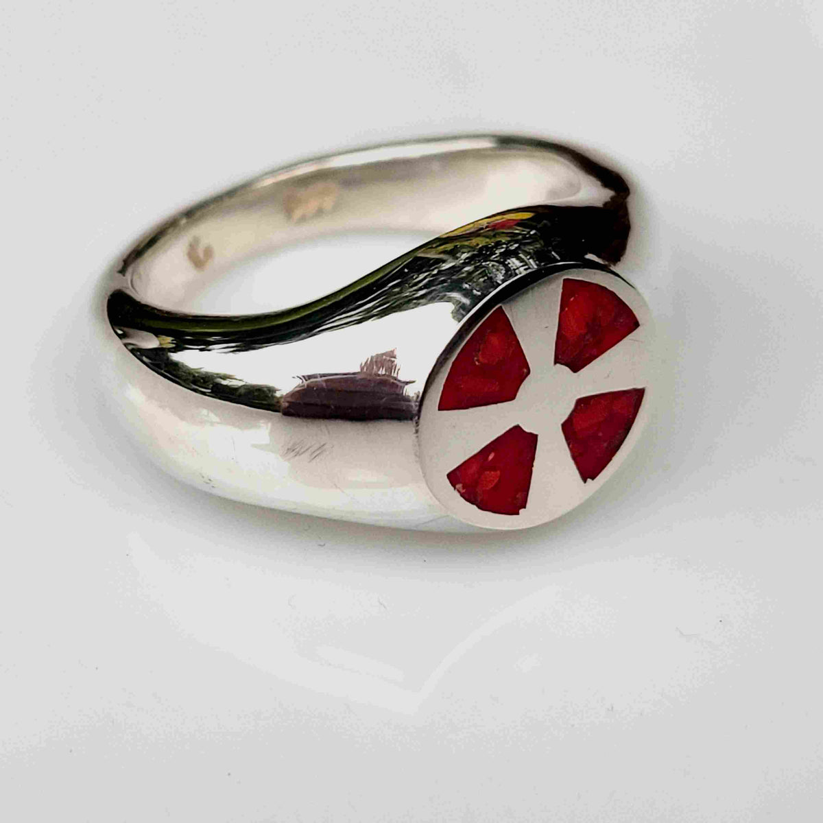 A polished silver ring featuring a circular face with a red and white cross-like emblem, photographed on a light gray reflective surface.