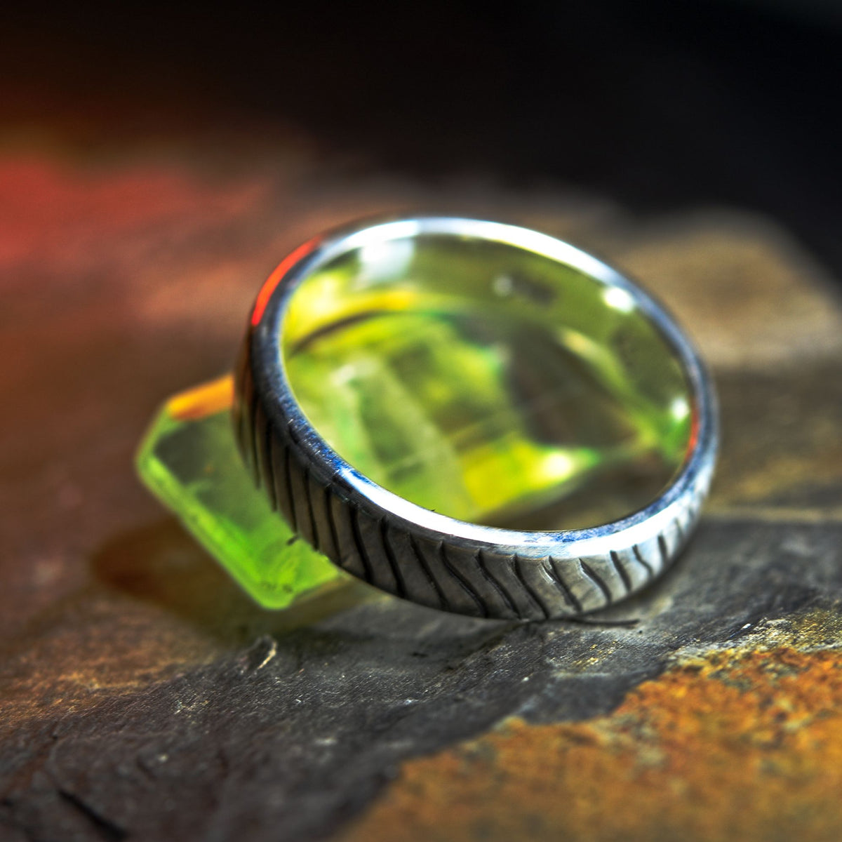 A silver ring with a textured, tread-like band and a clear bezel containing a vibrant green liquid, resting on a dark, rustic wooden surface.