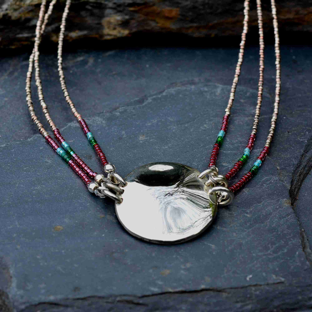 A layered necklace featuring a large, polished silver disc pendant suspended from three delicate strands of seed beads. The strands alternate between metallic silver, deep red, and green beads, resting on a dark slate surface.
