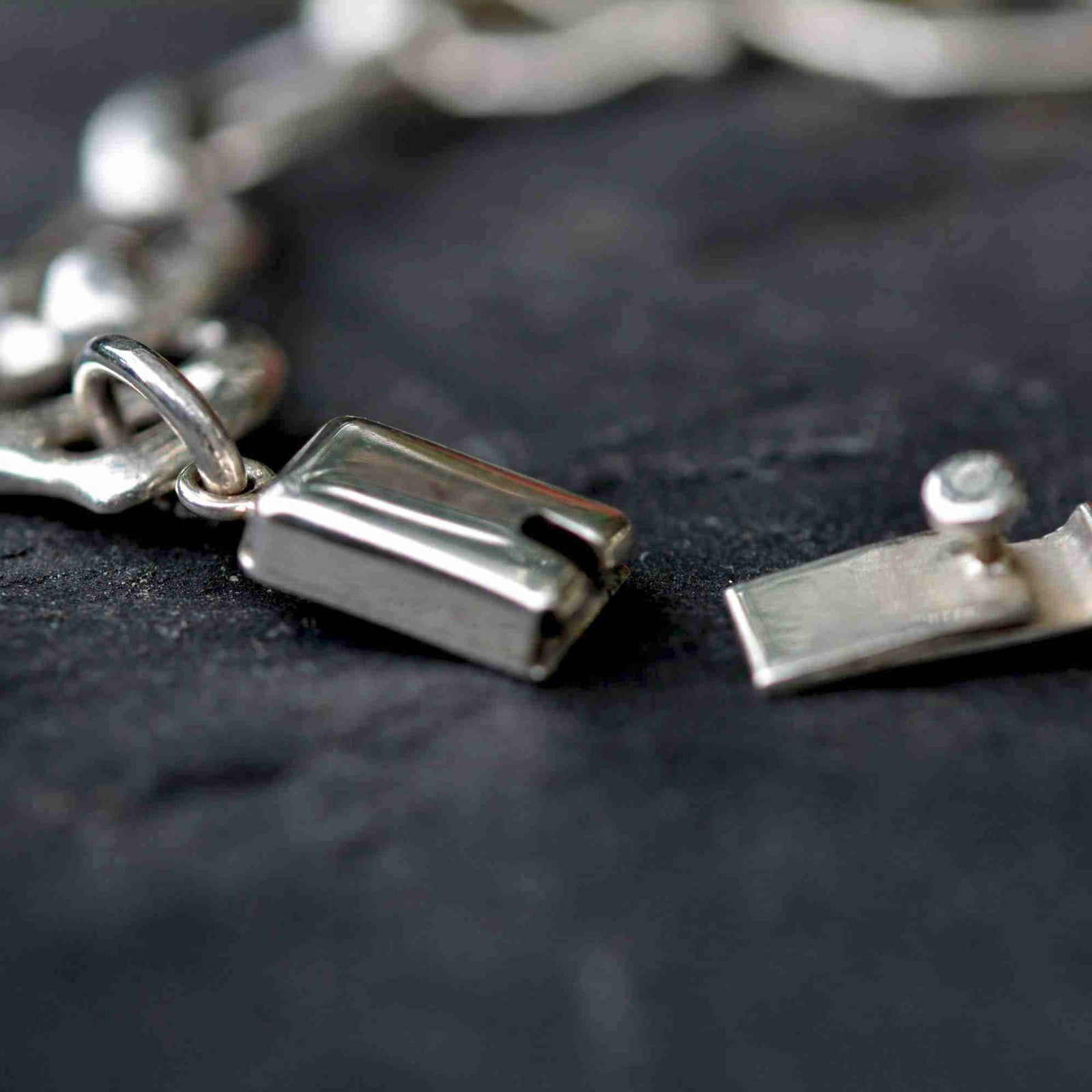 A close-up of a silver chain bracelet with a rectangular clasp and a small matching charm, resting on a dark textured surface.