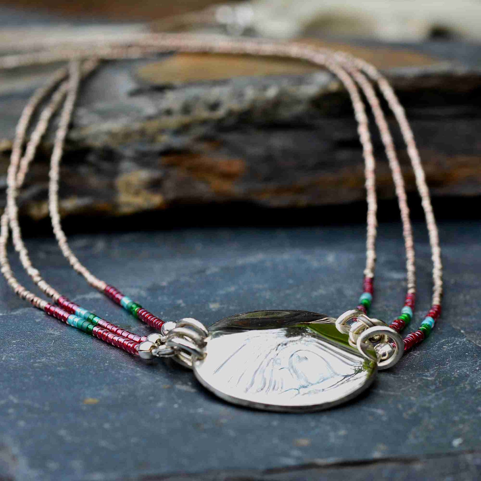 A silver-toned pendant necklace with a polished oval medallion, suspended from multiple delicate strands of small beads in red, green, turquoise, and cream, resting on a dark slate surface.