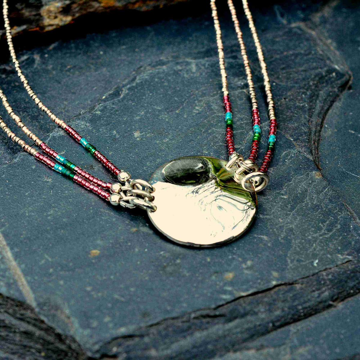 A close-up of a silver pendant necklace with a polished oval disc, hanging from multi-strand beaded cords in silver, red, and turquoise, displayed on a dark slate surface.