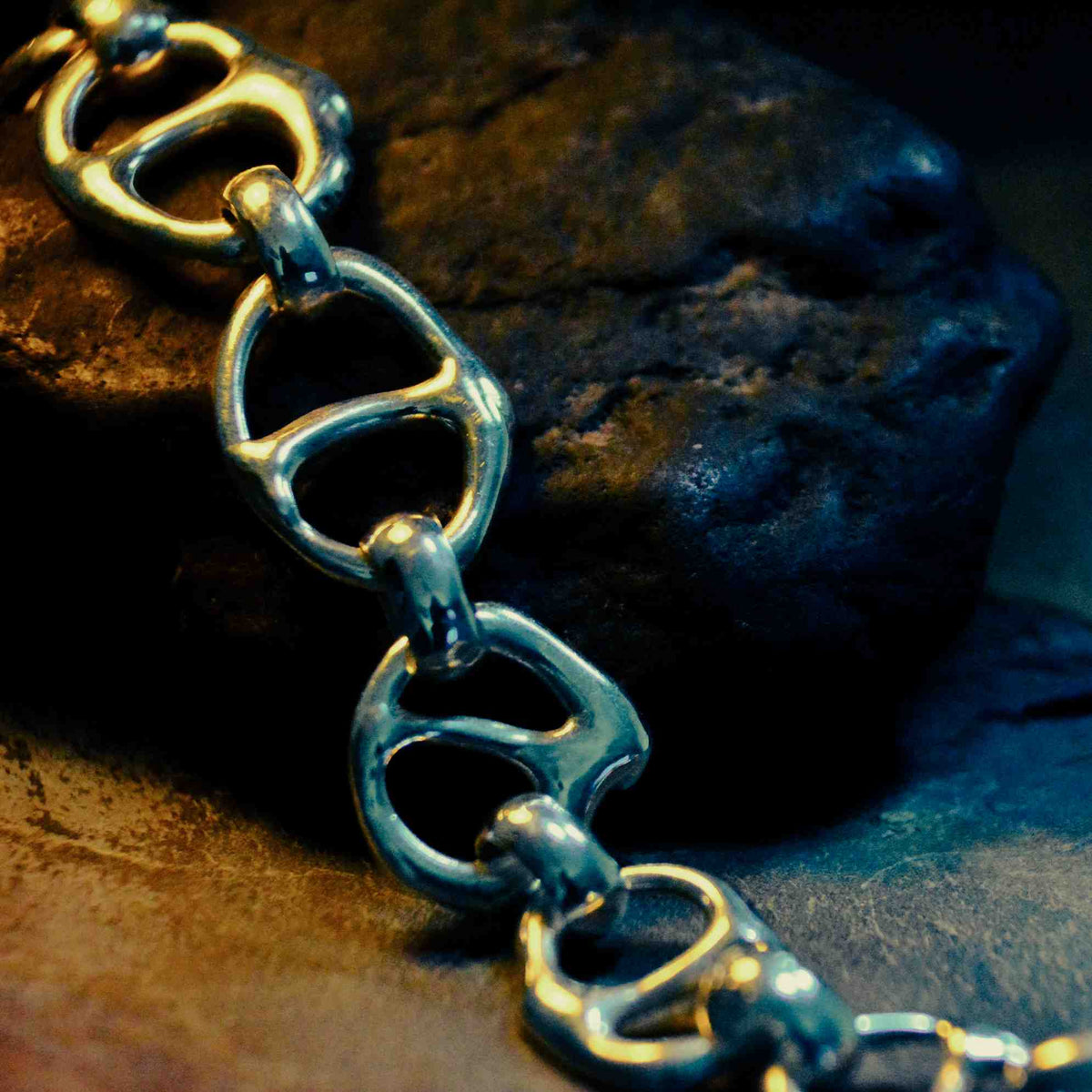 A close-up of a metallic link chain bracelet made from recycled soda cans, resting on a dark textured rock.