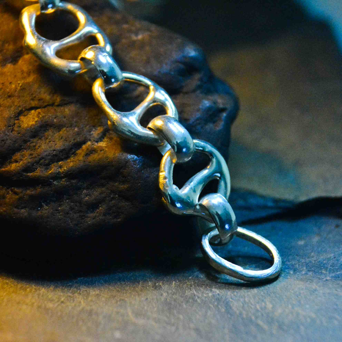 A close-up of a silver chain bracelet with interlocking oval links, draped over a dark textured rock on a dark surface.