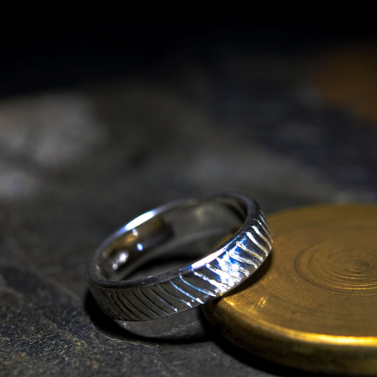 A silver band ring with a textured, ridged outer surface, resting on a dark stone surface next to a gold-colored coin.