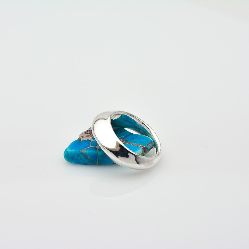 A polished silver ring with a curved, openwork design resting on a small piece of bright turquoise stone against a light gray background.