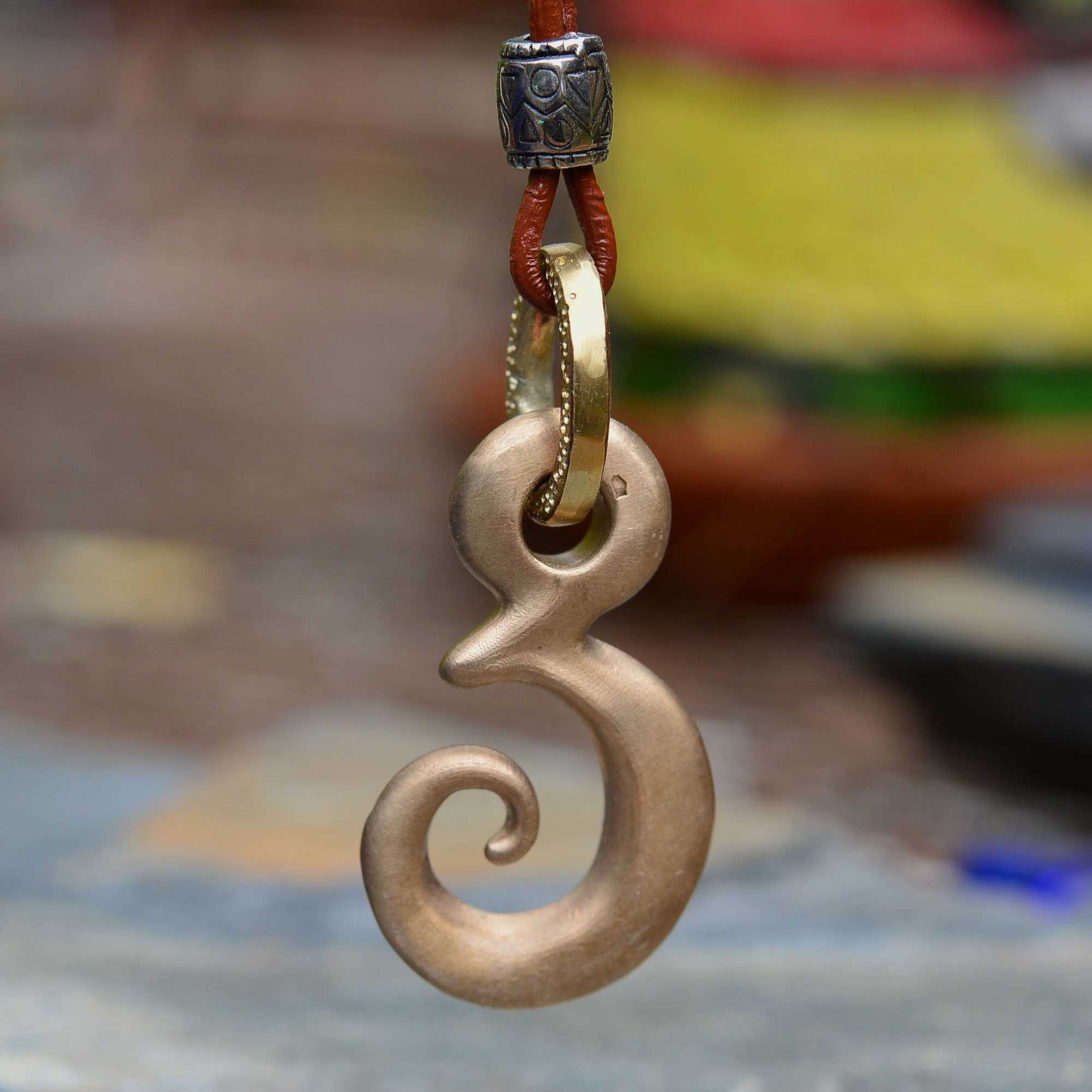 A polished silver-toned Maori tikanga pendant with a spiral design, hanging from a decorative chain against a blurred sepia-toned background.