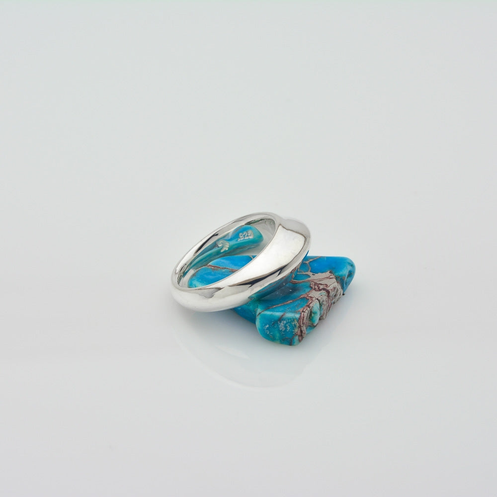 A silver ring with a curved band featuring inlaid turquoise and brown matrix, displayed on top of a larger turquoise stone against a clean white background.