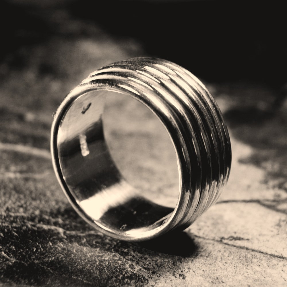 A silver-toned ring with a wide, horizontally grooved band, photographed on a dark textured surface with a shallow depth of field.