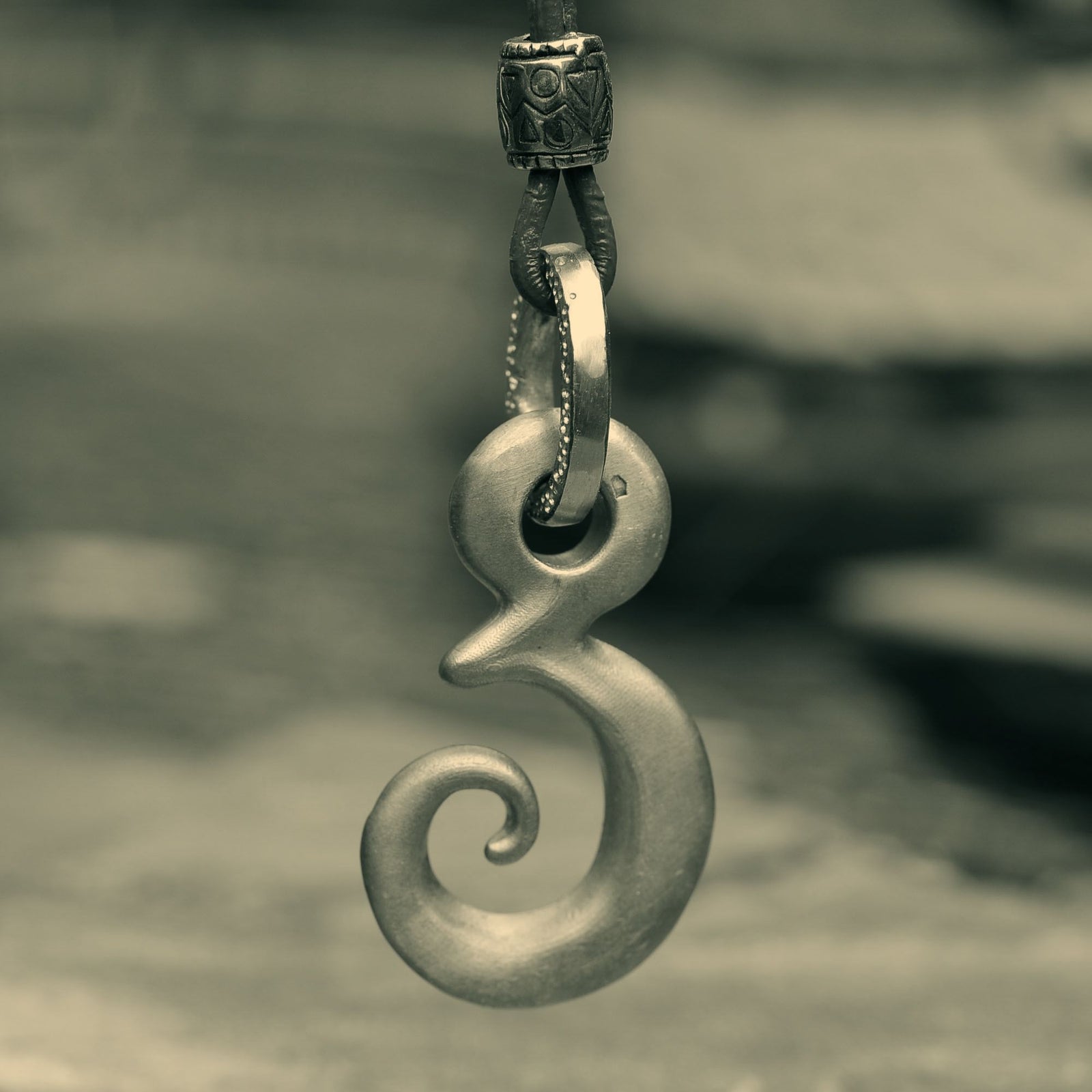 A polished silver-toned Maori tikanga pendant with a spiral design, hanging from a decorative chain against a blurred sepia-toned background.