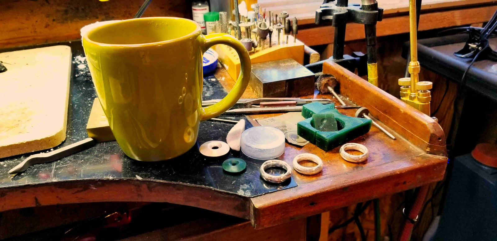 A yellow ceramic mug sits on a wooden workbench surrounded by small jewelry-making components and tools in a workshop setting.