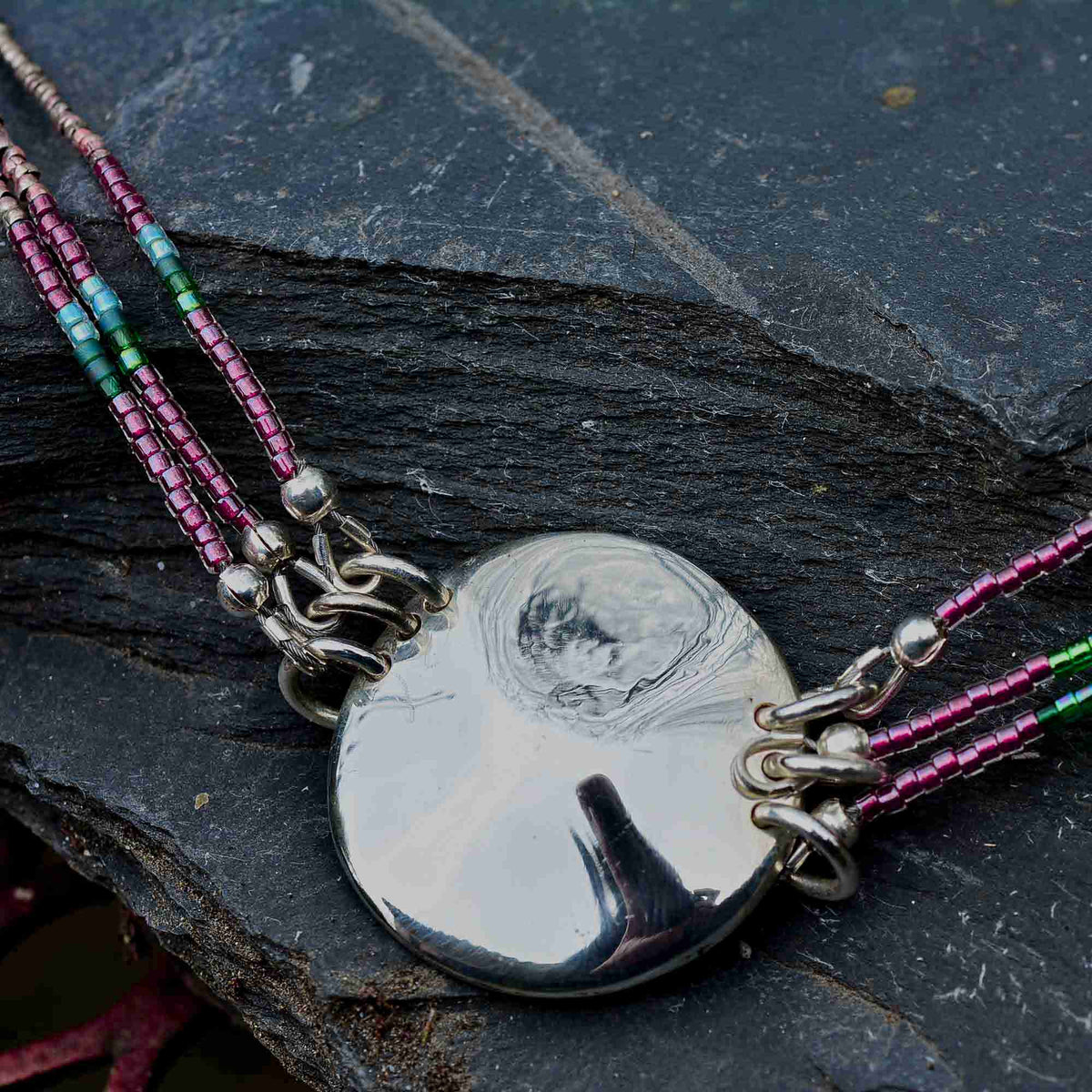 A close-up of a silver pendant necklace with a circular polished disc pendant and multi-strand beaded cords in pink, teal, and green, resting on a dark textured slate surface.