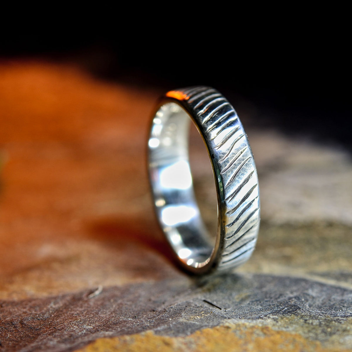 A silver ring with a textured, wavy engraved pattern, standing upright on a textured stone surface with a dark, out-of-focus background.