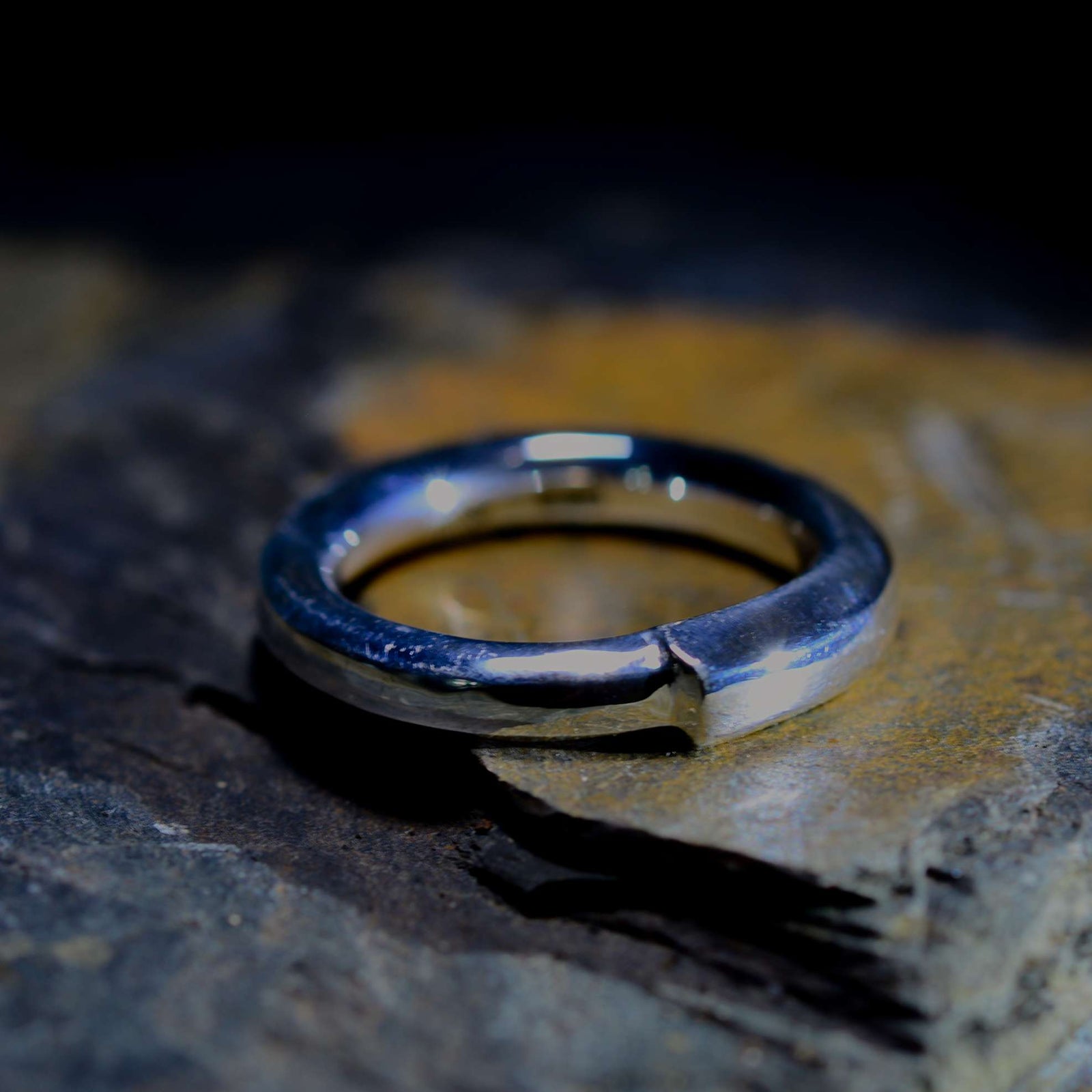 A textured silver band ring with a polished finish, resting on a dark, rough stone surface.