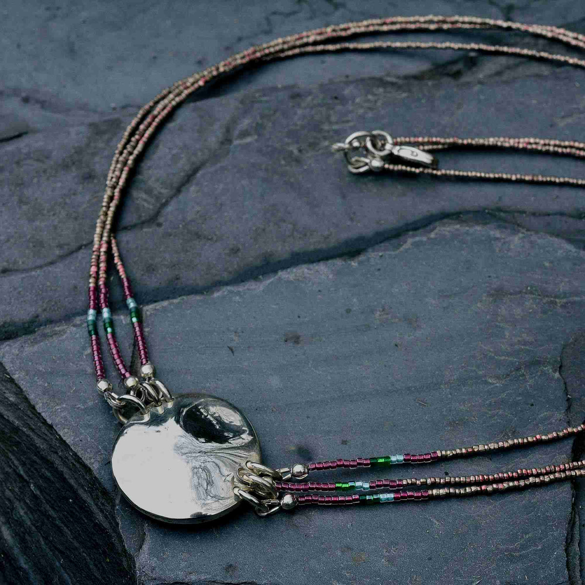 A beaded necklace with a polished silver disc pendant, laid out on a dark slate surface. The necklace features multiple strands of small beads in metallic rose gold, brown, and green, with the silver pendant hanging from silver clasps.
