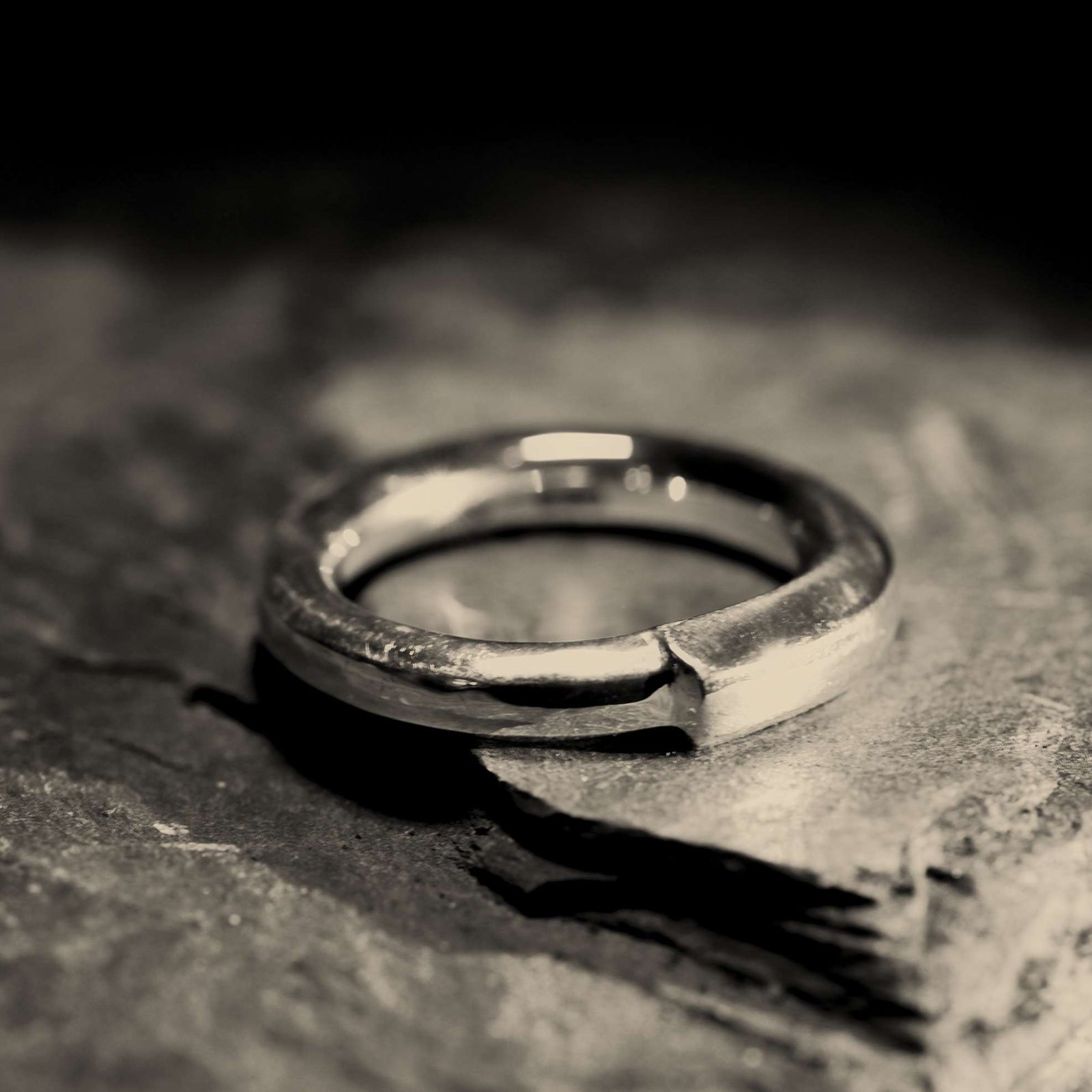 A textured silver band ring with a polished finish, resting on a dark, rough stone surface.
