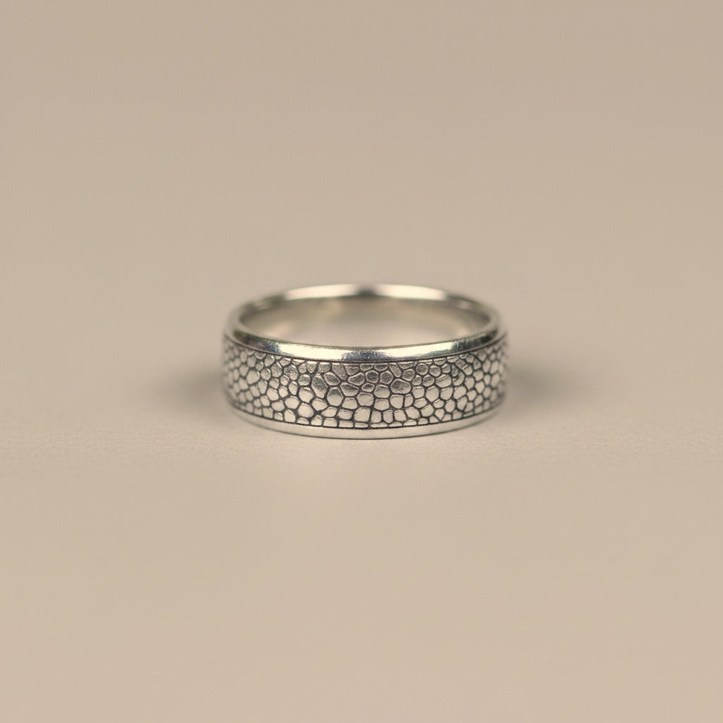 A silver band ring with a textured, raised honeycomb or scales-like pattern on the front, photographed on a neutral light gray background.